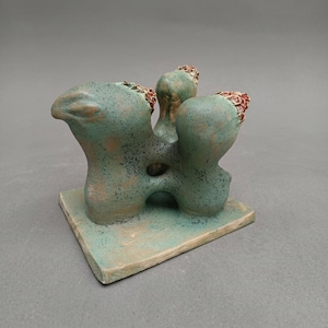 Puede incluir: Una escultura de cerámica que presenta tres formas redondeadas interconectadas de color verde azulado con superficies moteadas. Cada forma está rematada con una tapa texturizada de color marrón rojizo. Todo sobre una base cuadrada plana.