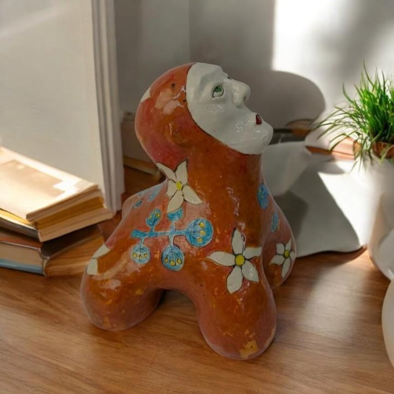May include: A ceramic sculpture of a person with a stylized face and floral patterns. The sculpture is painted in shades of orange, white, blue, and green.