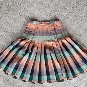 May include: A pleated skirt in a plaid pattern featuring shades of peach, teal, and navy. The skirt has a high waist and a flared silhouette. The fabric appears to be a woven material, suitable for a vintage or retro aesthetic.