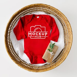 K&ouml;nnte beinhalten: Roter Langarm-Babybody mit einer wei&szlig;en Kamera-Grafik und dem Text "MOCKUP HUB". Der Body liegt in einem geflochtenen Korb.