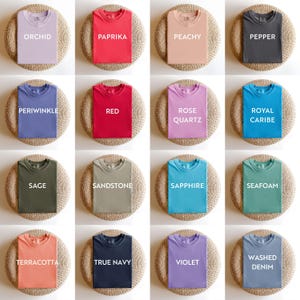 May include: A collection of folded t-shirts in various colors, including orchid, paprika, peachy, pepper, periwinkle, red, rose quartz, royal caribe, sage, sandstone, sapphire, seafoam, terracotta, true navy, violet, and washed denim. Each shirt has a color name printed on it.