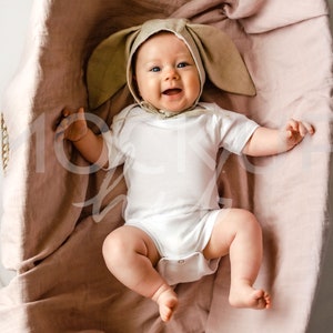 May include: A baby wearing a brown bunny hat and a white onesie is lying in a pink basket. The baby is smiling and looking at the camera.