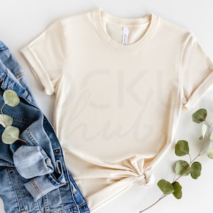 May include: A cream colored t-shirt with rolled up sleeves, a denim jacket, and a sprig of eucalyptus leaves on a white background.