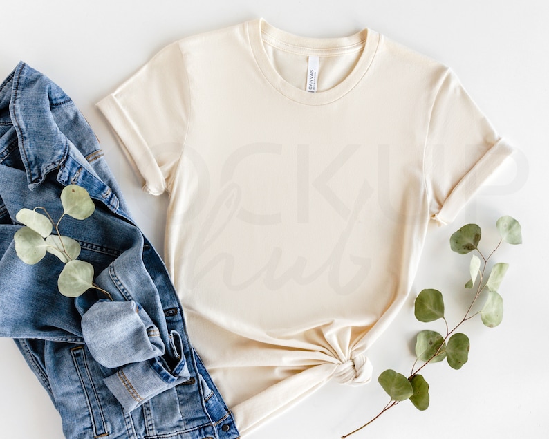 May include: A cream colored t-shirt with rolled up sleeves, a denim jacket, and a sprig of eucalyptus leaves on a white background.