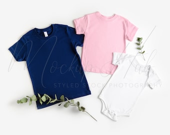 May include: Three blank t-shirts, one navy blue, one pink, and one white, laid flat on a white background with green sprigs of eucalyptus.