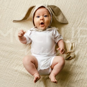 May include: A baby wearing a beige bunny ear hat and a white onesie is lying on a beige textured blanket. The baby is looking at the camera with a surprised expression.