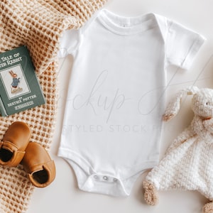 May include: A white baby onesie with short sleeves, a brown knitted blanket, a book titled "The Tale of Peter Rabbit" by Beatrix Potter, a pair of brown leather baby shoes, and a white plush bunny toy.