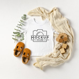 May include: White baby onesie with a black camera graphic and the text "MOCKUP - HUB". The onesie is laid flat on a white surface with a beige knitted blanket, a wooden toy car, and wooden blocks.