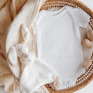 May include: A white baby onesie in a brown wicker basket, with a white crocheted stuffed animal and a beige muslin blanket.