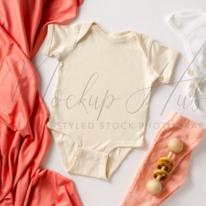 Op de afbeelding: Een witte baby romper met een koraal kleurige baby muts, broek en deken. Een houten rammelaar is ook inbegrepen.