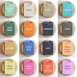 May include: A collection of folded t-shirts in various colors, including green, ivory, khaki, and blue. Each shirt displays a color name in white text, such as "Island Reef" and "Lagoon". The shirts are arranged on round, woven mats.