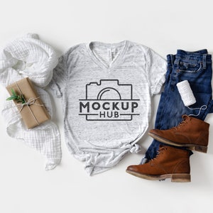 May include: A white heather grey V-neck t-shirt with a camera graphic and the text "MOCKUP HUB". The t-shirt is laid flat on a white surface with a brown gift box, a white scarf, blue jeans, and brown boots.