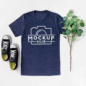 May include: A navy blue t-shirt with a white camera graphic and the text "MOCKUP HUB" printed on the front. The t-shirt is laid flat on a white surface with a pair of black sneakers and a green plant sprig.