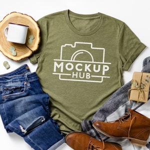 Pode incluir: Uma camiseta verde com um gráfico de câmera branco e o texto "MOCKUP HUB". A camiseta está colocada sobre uma superfície branca com jeans azul, botas marrons, um cobertor branco e uma pequena bandeja de madeira com uma caneca branca e folhas verdes.