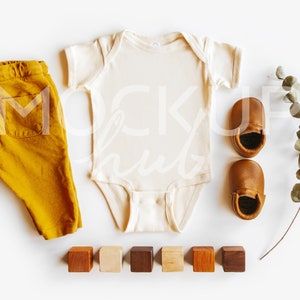 May include: A white baby onesie, mustard yellow pants, brown leather baby shoes, a sprig of eucalyptus, and six wooden blocks in various shades of brown.