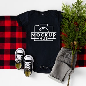May include: A black baby onesie with a white camera graphic and the text "MOCKUP HUB". The onesie is laid out on a red and black plaid blanket with a pair of black and white sneakers and a sprig of green foliage.