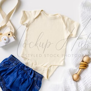 May include: A white onesie with a wooden rattle, a blue denim nappy cover, a wooden toy camera, and a white muslin swaddle blanket.