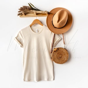 Può includere: Una maglietta beige su un appendiabiti con un cappello marrone e una borsa rotonda intrecciata appesa a un muro bianco.