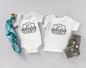 May include: Two white baby onesies with short sleeves. The onesies have a black camera graphic and the text "MOCKUP HUB". One onesie is a bodysuit and the other is a t-shirt. A pair of gray pants with three colorful buttons is also included.