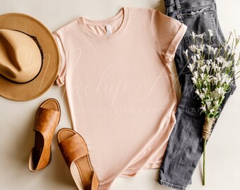 May include: A flat lay featuring a light pink t-shirt, a brown hat, brown sandals, and gray jeans. A bouquet of white flowers tied with twine rests next to the jeans. The t-shirt is a crew neck style.