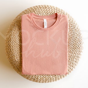 May include: A folded peach-colored t-shirt on a woven straw mat. The t-shirt has a round neckline and a tag that reads "Canvas" in white.