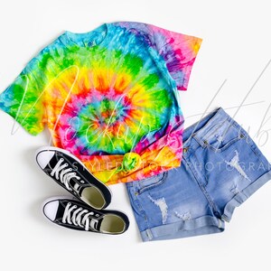 May include: A tie-dye t-shirt with a rainbow swirl pattern, blue denim shorts with distressed details, and black canvas sneakers.