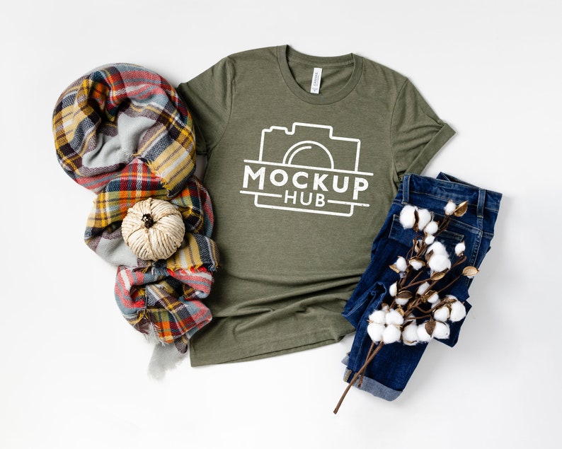 May include: A green t-shirt with a white camera graphic and the text "MOCKUP HUB" on the front. The t-shirt is laid flat on a white surface with a plaid scarf, a small white pumpkin, and a pair of blue jeans with a cotton branch.