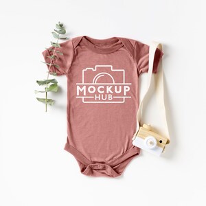 Pode incluir: Um macacão de bebê malva com um gráfico de câmera branca e o texto "MOCKUP HUB" na frente. Uma pequena câmera de brinquedo de madeira com uma lente branca em um fundo branco.