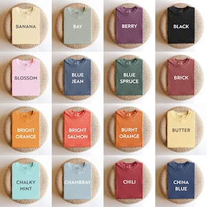May include: A collection of sixteen t-shirts in various colors, including yellow, green, purple, and black. Each shirt is displayed on a round, woven surface. The shirts have text labels such as "Banana," "Bay," and "Berry."