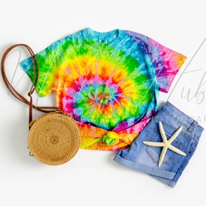 May include: A tie-dye t-shirt with a rainbow swirl pattern, blue denim shorts, and a round woven straw bag with a brown leather strap.