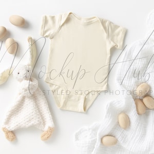 May include: A white baby onesie with a white bunny rattle and wooden eggs on a white background. The onesie is blank and ready for personalisation.