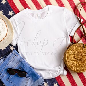 Puede incluir: Una camiseta blanca, unos pantalones cortos de mezclilla azul, un bolso de paja con una correa marr&oacute;n y un sombrero de sol blanco sobre una tela estampada en rojo, blanco y azul.