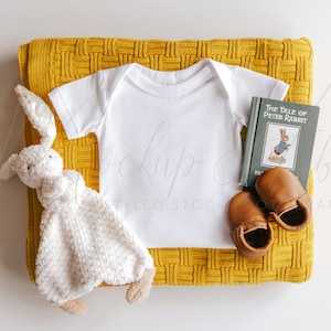 May include: A white baby onesie on a yellow knitted blanket with a white bunny lovey, a book titled "The Tale of Peter Rabbit", and brown leather baby shoes.