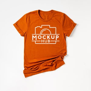 May include: A folded orange t-shirt with a white camera graphic and the text "MOCKUP HUB" printed on the front.