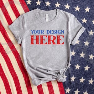May include: A heather gray t-shirt with the text "YOUR DESIGN HERE" in blue and red. The shirt is displayed on a background of a US flag.