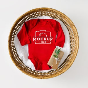 K&ouml;nnte beinhalten: Roter lang&auml;rmliger Baby-Strampler mit wei&szlig;em Text "MOCKUP HUB" und einem Kamerasymbol. Der Strampler liegt in einem geflochtenen Korb mit einer kleinen Geschenkbox.