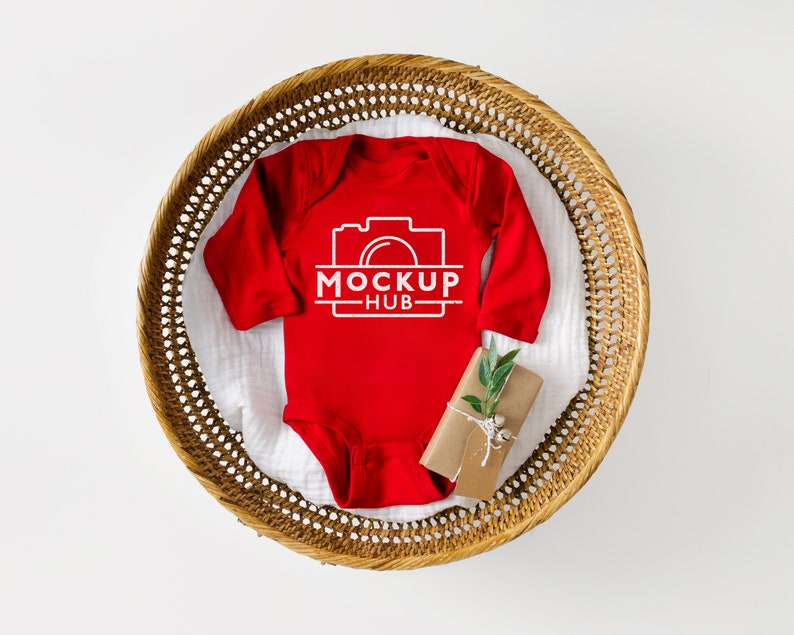 K&ouml;nnte beinhalten: Roter lang&auml;rmliger Baby-Strampler mit wei&szlig;em Text "MOCKUP HUB" und einem Kamerasymbol. Der Strampler liegt in einem geflochtenen Korb mit einer kleinen Geschenkbox.