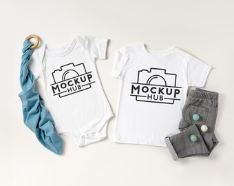 May include: Two white baby shirts with short sleeves, one a bodysuit and one a t-shirt, both featuring a black camera graphic and the text "MOCKUP HUB". A blue fabric swaddle and gray pants with white and blue buttons are also included.