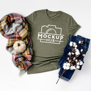 May include: A green t-shirt with a white camera graphic and the text "MOCKUP HUB" printed on the front. The t-shirt is laid flat on a white surface with a plaid scarf, a small white pumpkin, and a pair of blue jeans with a cotton branch.