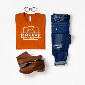 May include: A flat lay of a burnt orange t-shirt with a white camera graphic and the text "MOCKUP HUB" on it.  The t-shirt is laid out on a white surface with a pair of brown leather boots, a pair of blue jeans, and a pair of brown tortoise shell glasses.