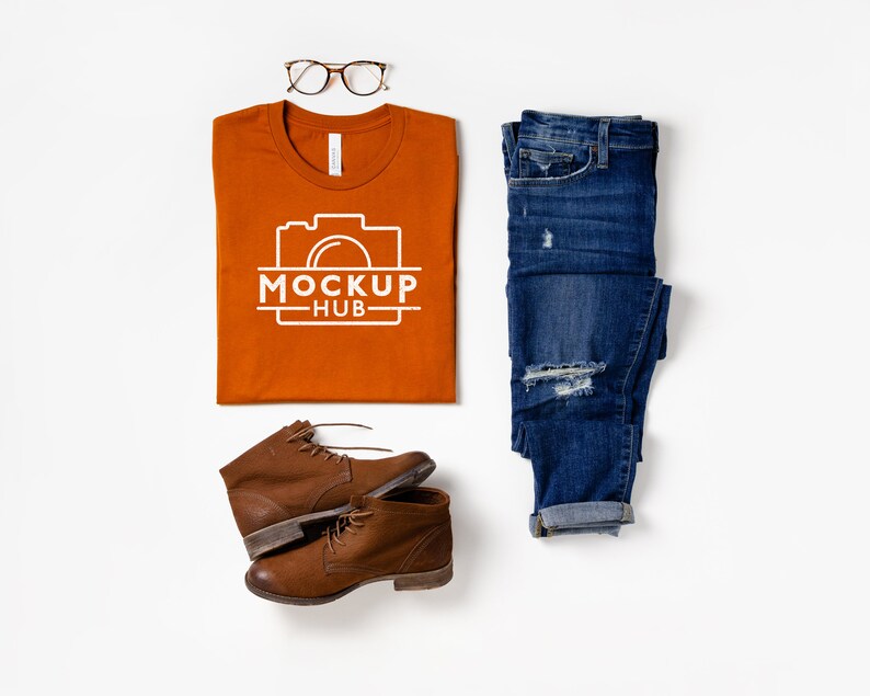 May include: A flat lay of a burnt orange t-shirt with a white camera graphic and the text "MOCKUP HUB" on it.  The t-shirt is laid out on a white surface with a pair of brown leather boots, a pair of blue jeans, and a pair of brown tortoise shell glasses.