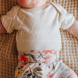 May include: A baby wearing a white short-sleeved onesie and floral print shorts. The baby is lying on a beige textured blanket.