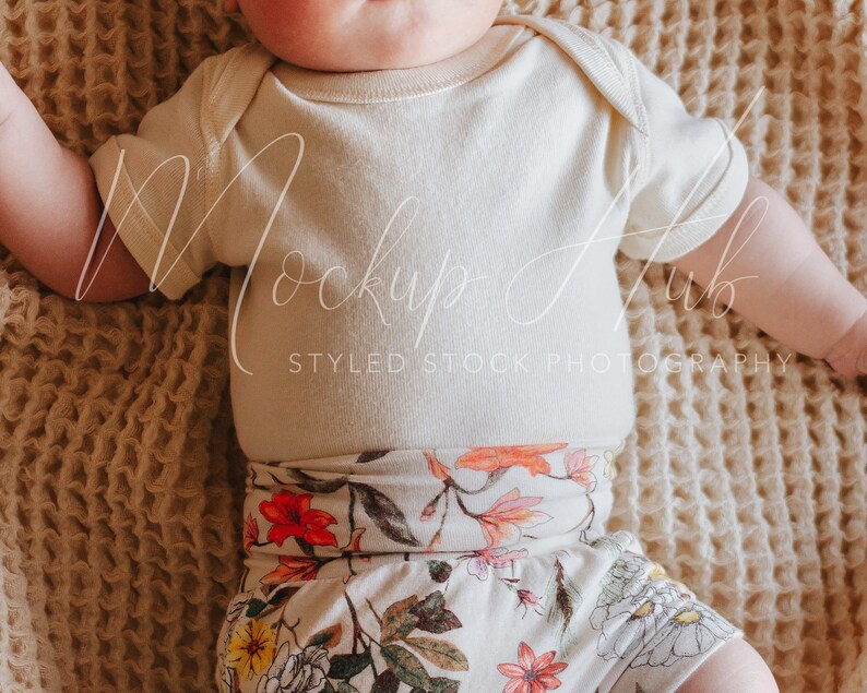May include: A baby wearing a white short-sleeved onesie and floral print shorts. The baby is lying on a beige textured blanket.
