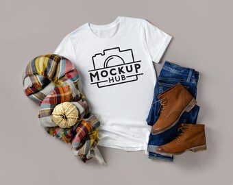 May include: White t-shirt with a black camera graphic and the text "MOCKUP HUB". The t-shirt is laid flat on a gray surface with a plaid scarf, blue jeans, and brown boots.