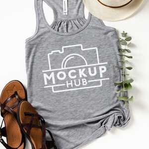 May include: A heather gray tank top with the text "MOCKUP HUB" in white, along with a camera graphic. The image also includes brown sandals, a straw hat, and a sprig of green leaves, all arranged on a white surface.