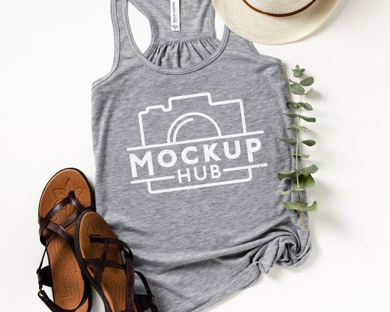 May include: A heather gray tank top with the text "MOCKUP HUB" in white, along with a camera graphic. The image also includes brown sandals, a straw hat, and a sprig of green leaves, all arranged on a white surface.