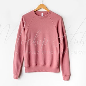 以下が含まれることがあります： 白の背景に、木製のハンガーにかけられたダスティローズ色のクルーネック長袖スウェットシャツ。