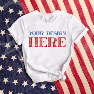 May include: White t-shirt with the text "YOUR DESIGN HERE" in blue and red. The shirt is knotted at the bottom and is displayed on a background of the American flag.