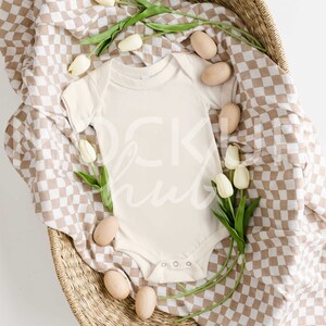 May include: A white baby onesie in a wicker basket with a brown and white checked blanket. White tulips and wooden eggs are scattered around the onesie.
