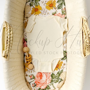 May include: A white baby onesie in a wicker basket with a floral patterned liner. A wooden rattle is next to the onesie.